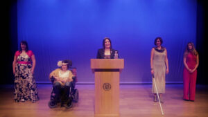 4 mujeres con discapacidad en un escenario y una mujer de presentadora en un atril