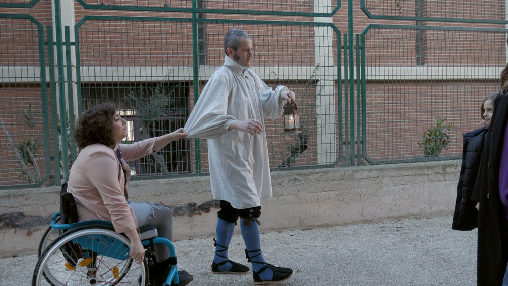 mujer con discapacidad en silla de ruedas caminando junto a un hombre vestido de fantasma con un candil por la calle. La mujer le agarra del brazo.