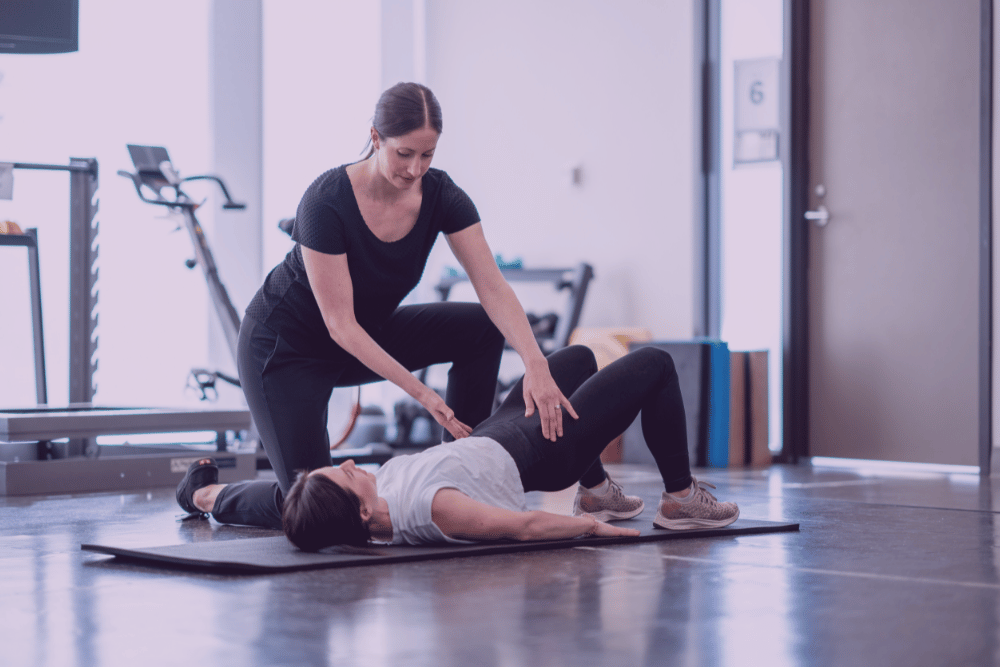 Fisioterapeuta ayudando a realizar una postura a otra mujer para fortalecer el suelo pélvico en un gimnasio sobre una esterilla